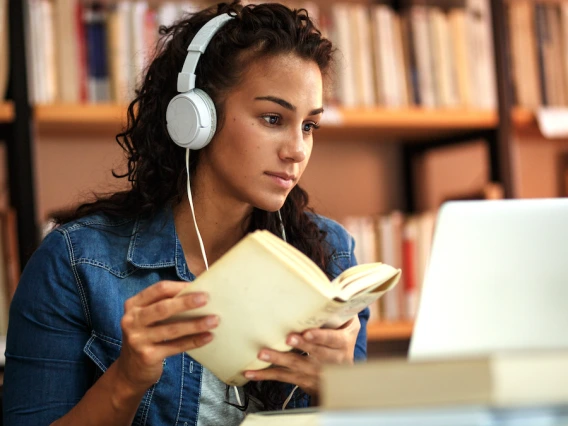 Woman wearing headphones with book in front of laptop in library