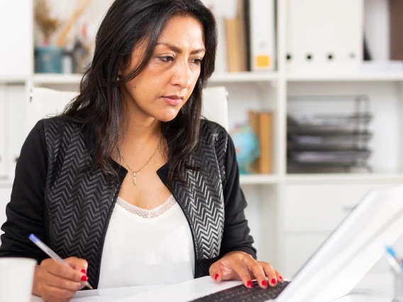 A woman with a laptop and a pencil in one hand, taking notes on paper