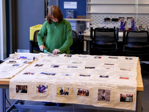 A student in a green sweater works on a fabric project with numerous photos and text on a table in the Maker Studio.