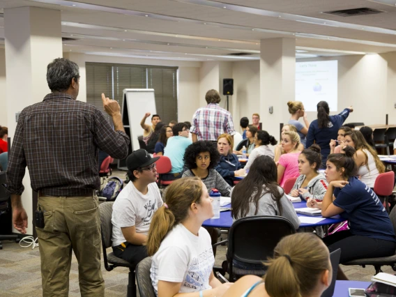 Instructor standing among students working in groups at several tables