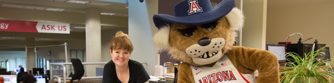 Library visitor stands with Wilbur Wildcat, the University of Arizona mascot, at the Ask Us desk in the library.