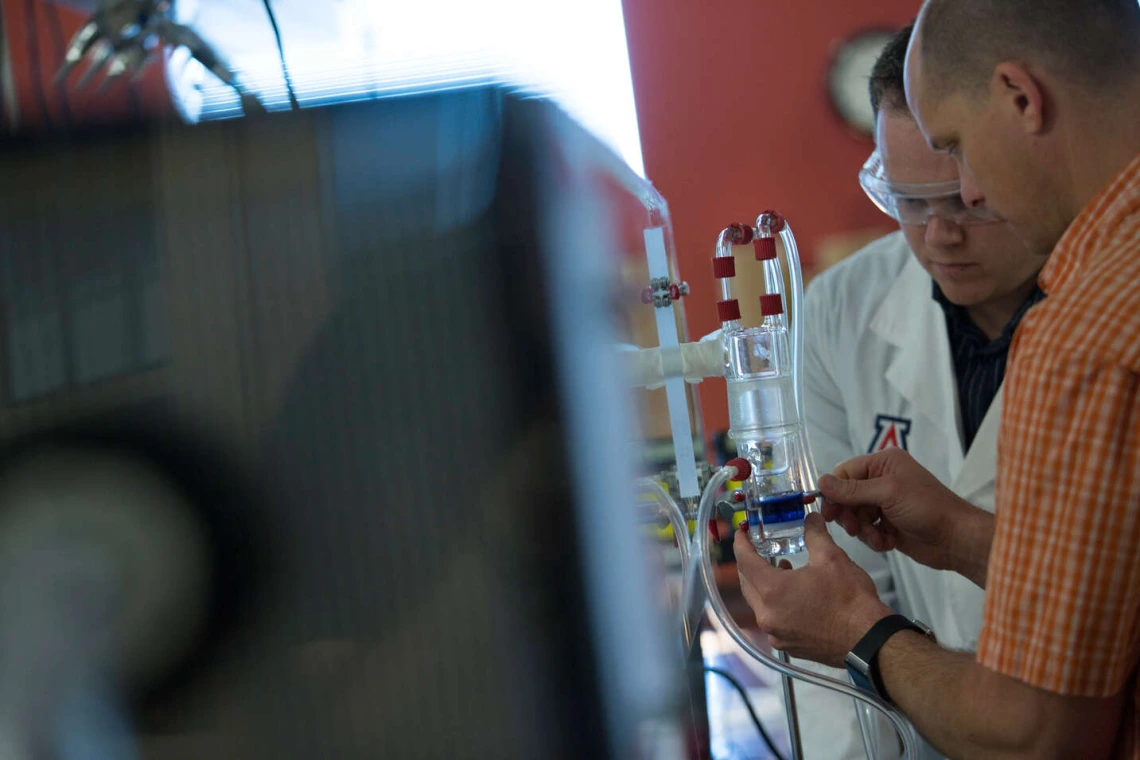 Researcher in white lab coat working in lab with person in orange shirt