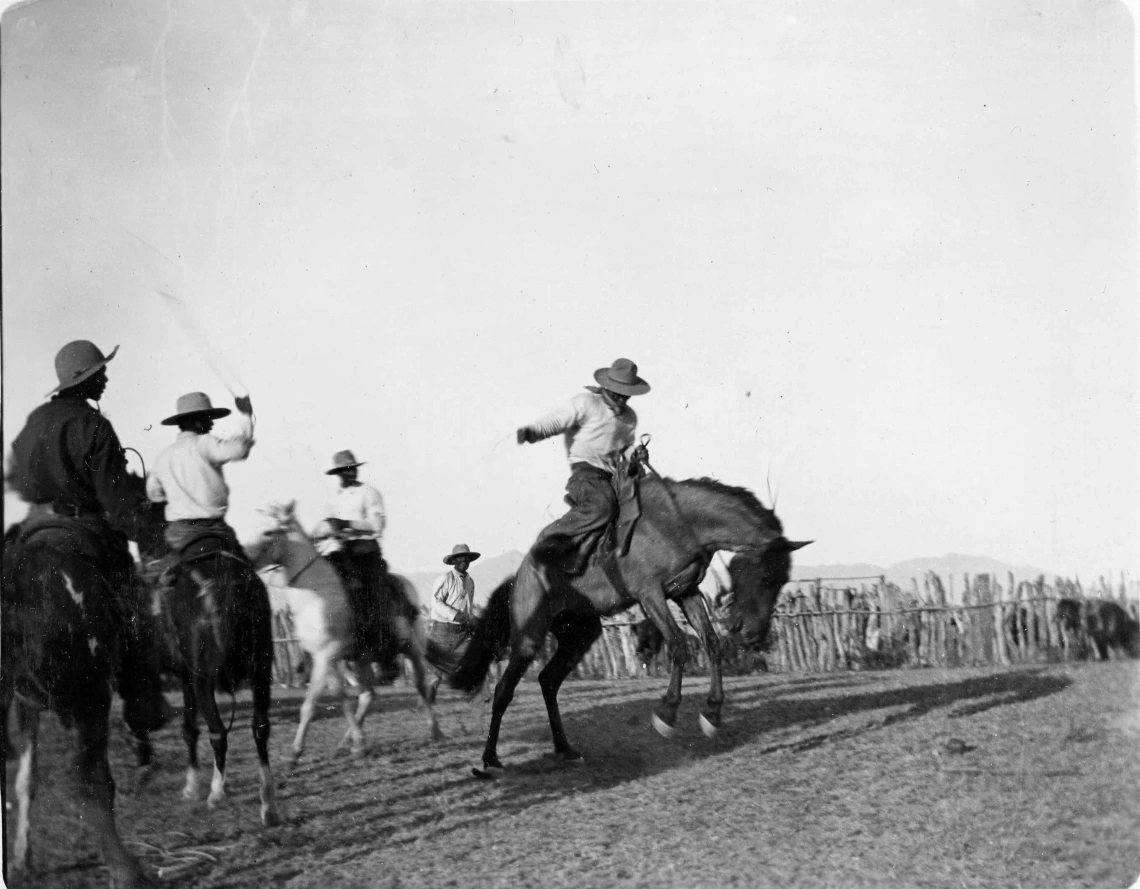 A black and white photograph depicting a man riding a bucking horse at Empire Ranch. Other men on horseback look on. 