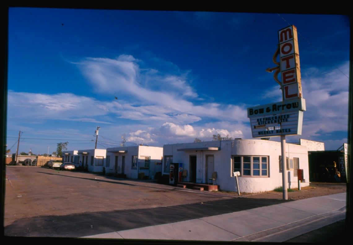 Landscape photo of a motor court. There are four white-walled motor court motel buildings behind a marquee street sign labelled Bow & Arrow Motel.