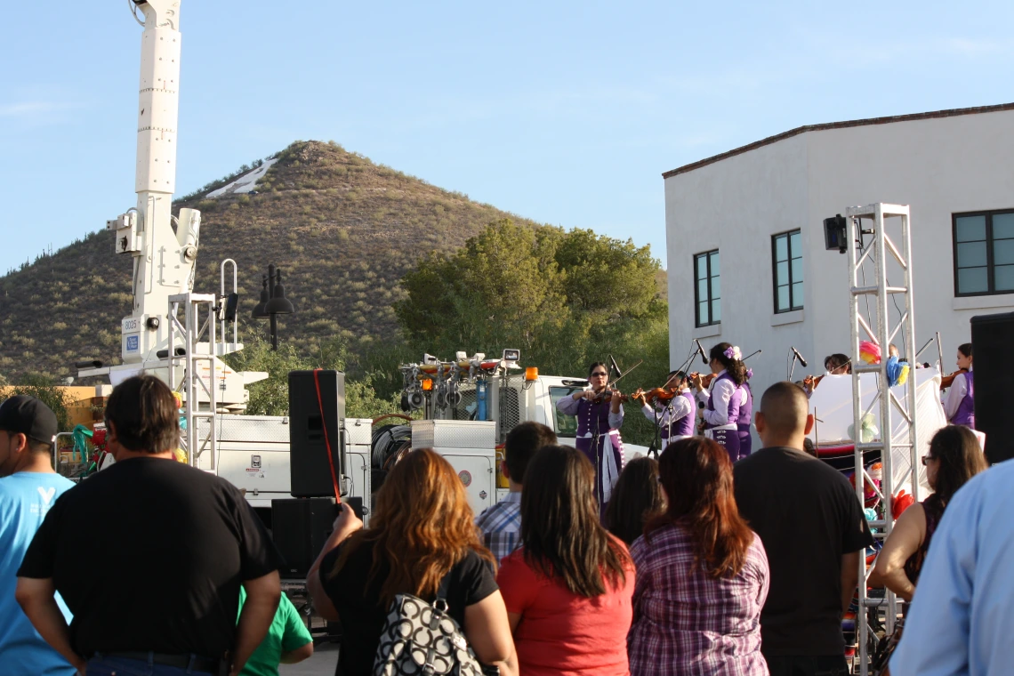 Landscape photograph. A small crowd faces an outdoor stage away from the photographer. Mariachi violin performers in purple charro suits play on the stage. “A” mountain and a white-walled house can be seen in the background.