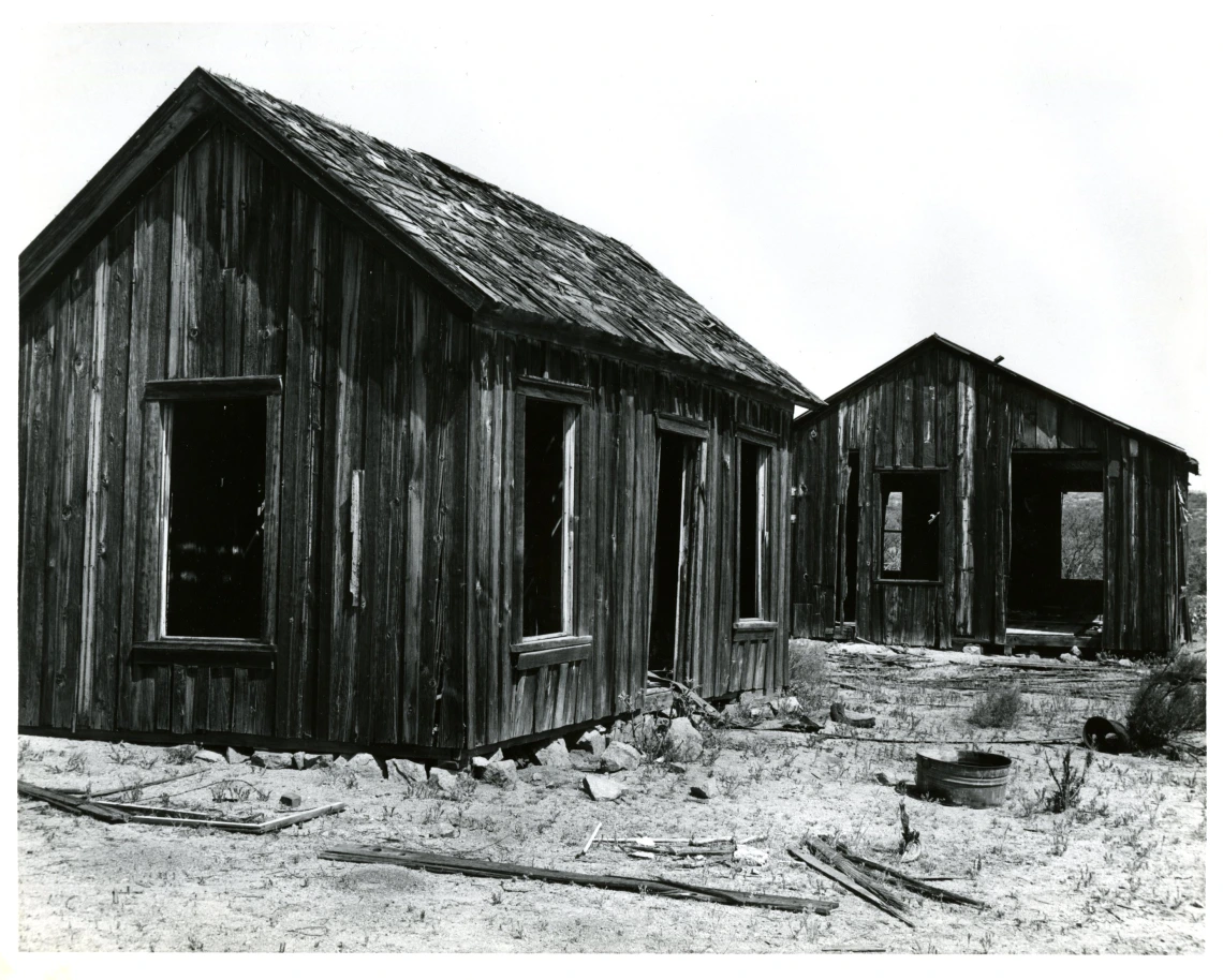 Two abandoned houses with wooden paneling taken in black and white. The houses are in states of disrepair.