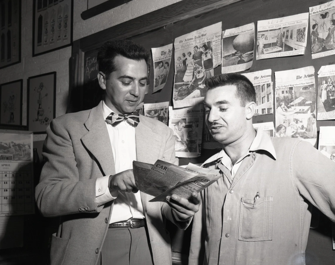 Black and white photo of two men inspecting newspaper clippings. Sam Levitz is at left and wears a sport coat and bow tie.