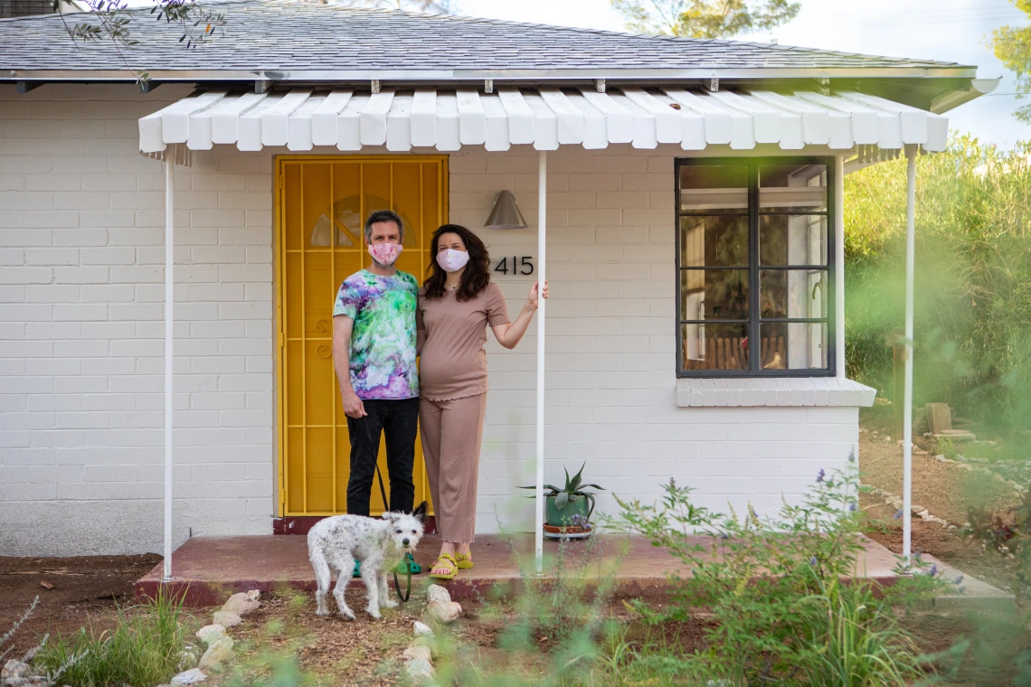 David Fenster, Laura Copelin, and Buddy, a black and white terrier, stand on the porch outside their home. David and Laura wear fabric masks.