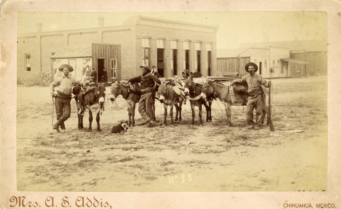 A sepia cabinet card photo of three men with a group of burros or mules posing on a dirt street in Phoenix, Arizona circa 1887.