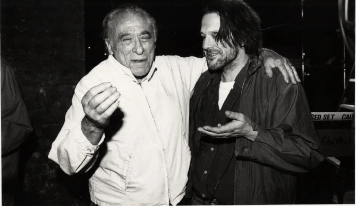 In a black and white image, writer Charles Bukowski has his arm around actor Mickey Rourke. The two are gesturing and smiling.