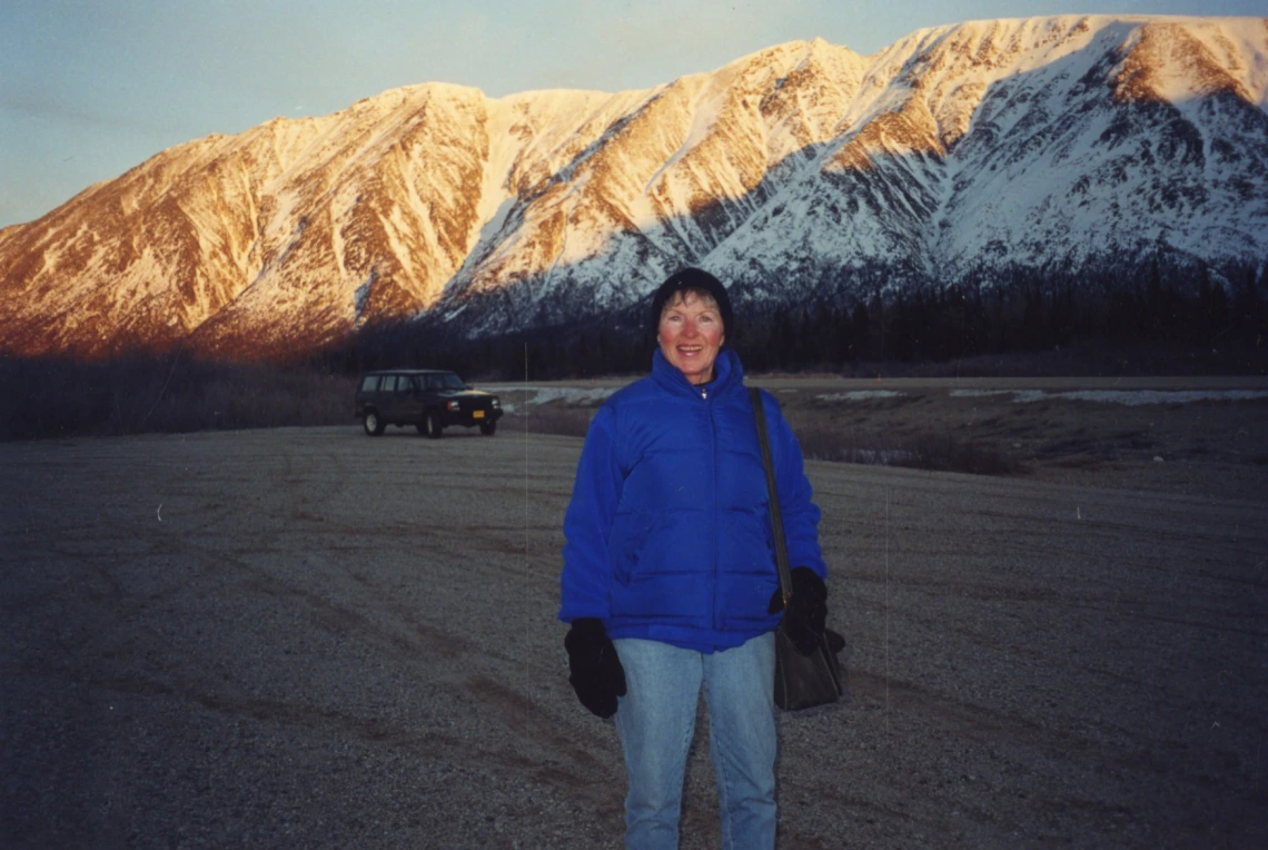 Color photograph of author Elizabeth Gunn. She is smiling and wears a blue winter coat and black mittens. Behind her is a Jeep and snow covered mountains.