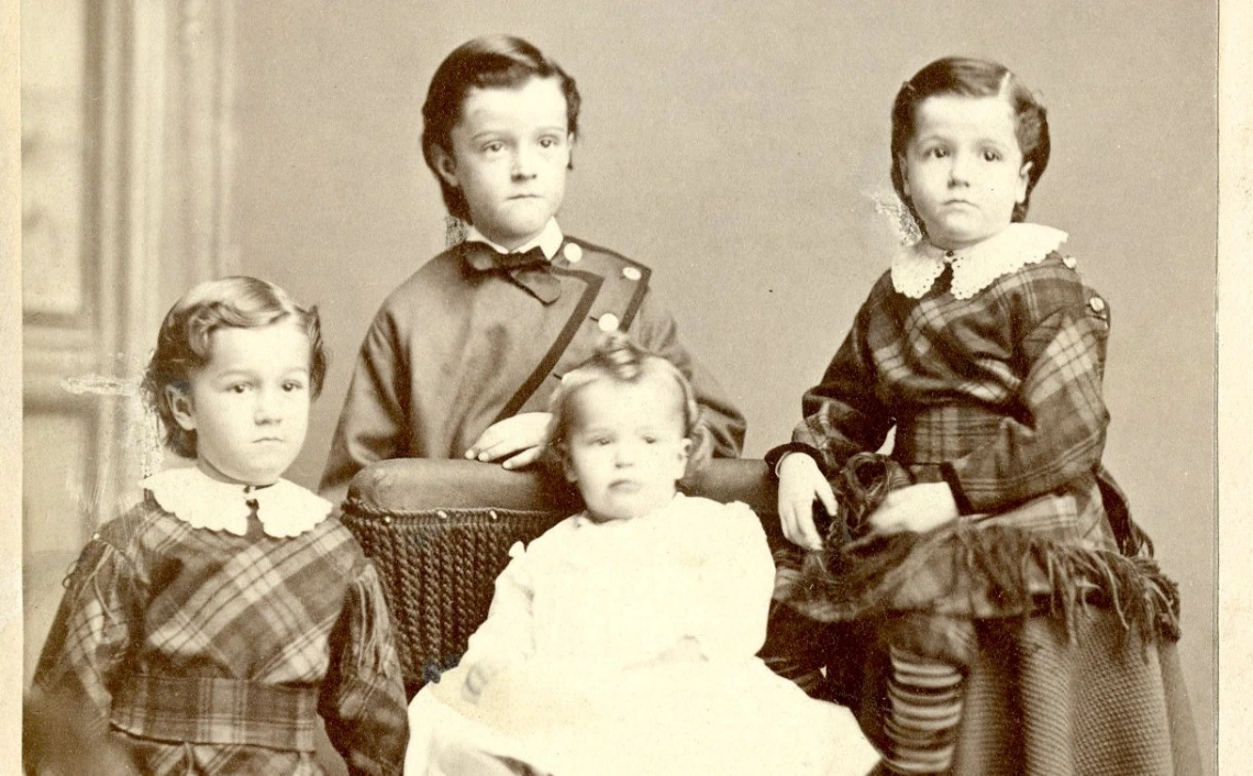 Black and white portrait of four children from the Hodge family. Three are gathered around a chair, where an infant in white is seated.