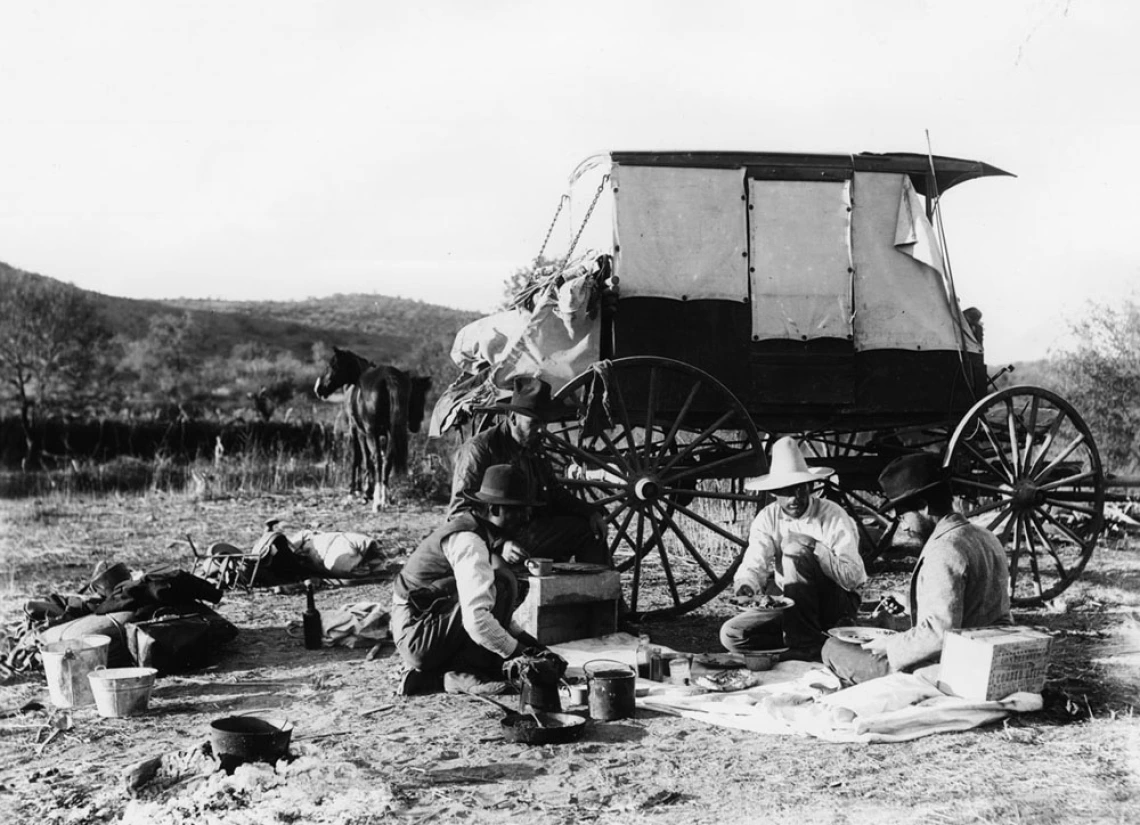 4 men eating a meal. A wagon and their horses are in the background.