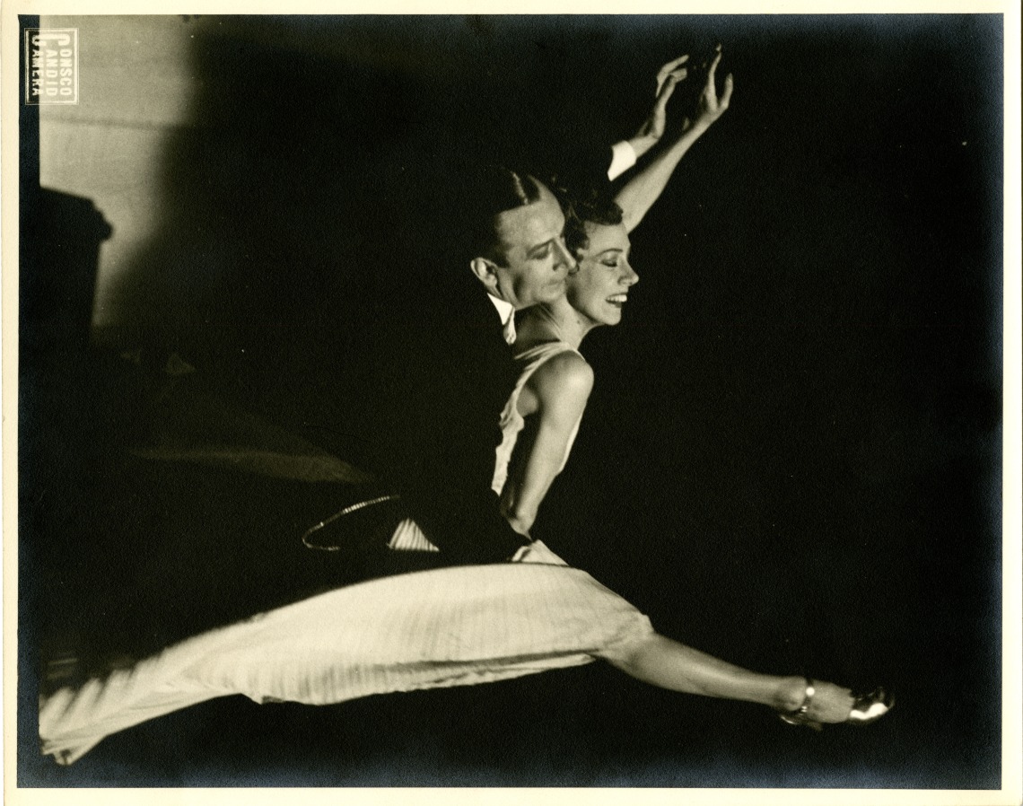 Black and white photograph of Billee Maye with partner Harry Roye performing a dance together. Both dancers are mid-leap with their left arms raised.