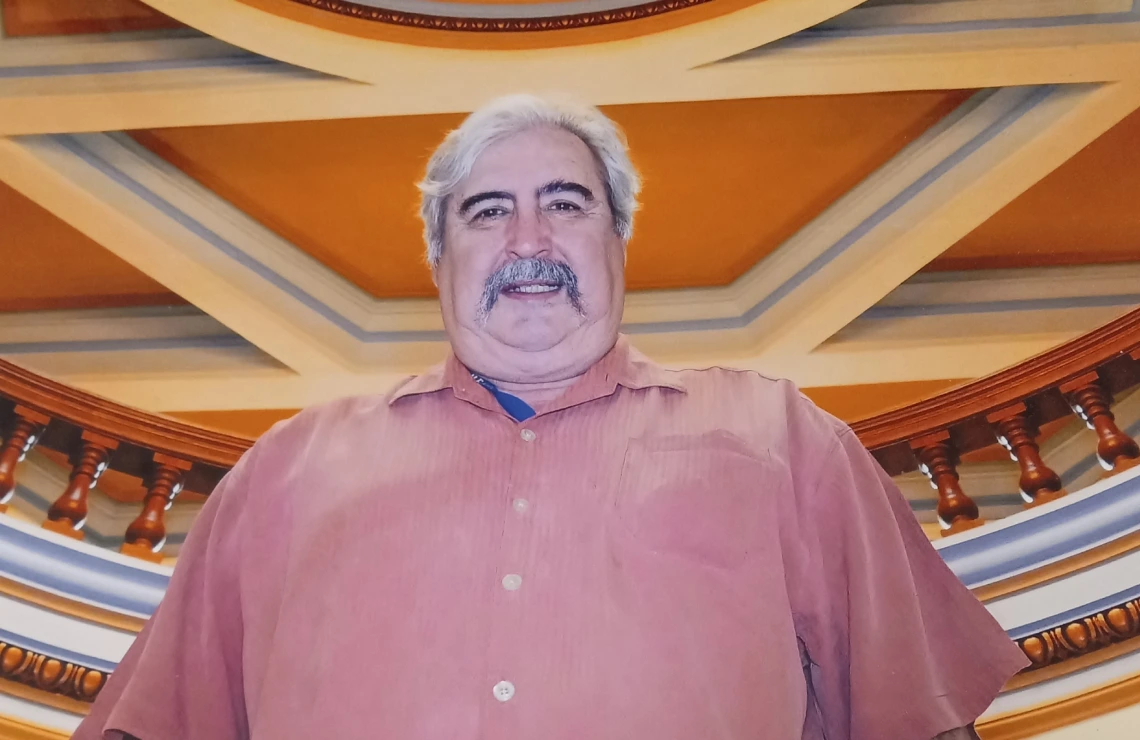 Low angle portrait of Dr. Jack August, wearing a rust colored button down shirt and framed by a neoclassical ceiling