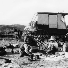 4 men eating a meal. A wagon and their horses are in the background.