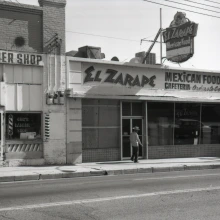In this street view, El Zarape, a Mexican food and cafeteria restaurant, is featured. Above the restaurant is a neon sign advertising the establishment.