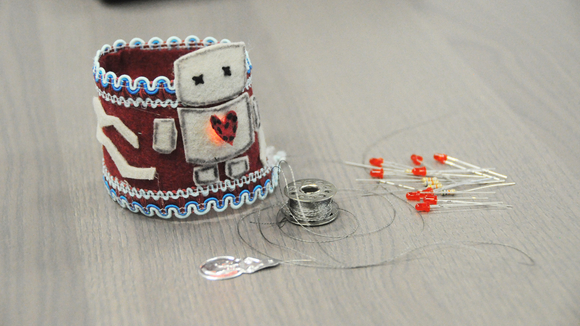 Photo showing a wearable bracelet designed with a felt robot and light up red heart, plus thread and pins